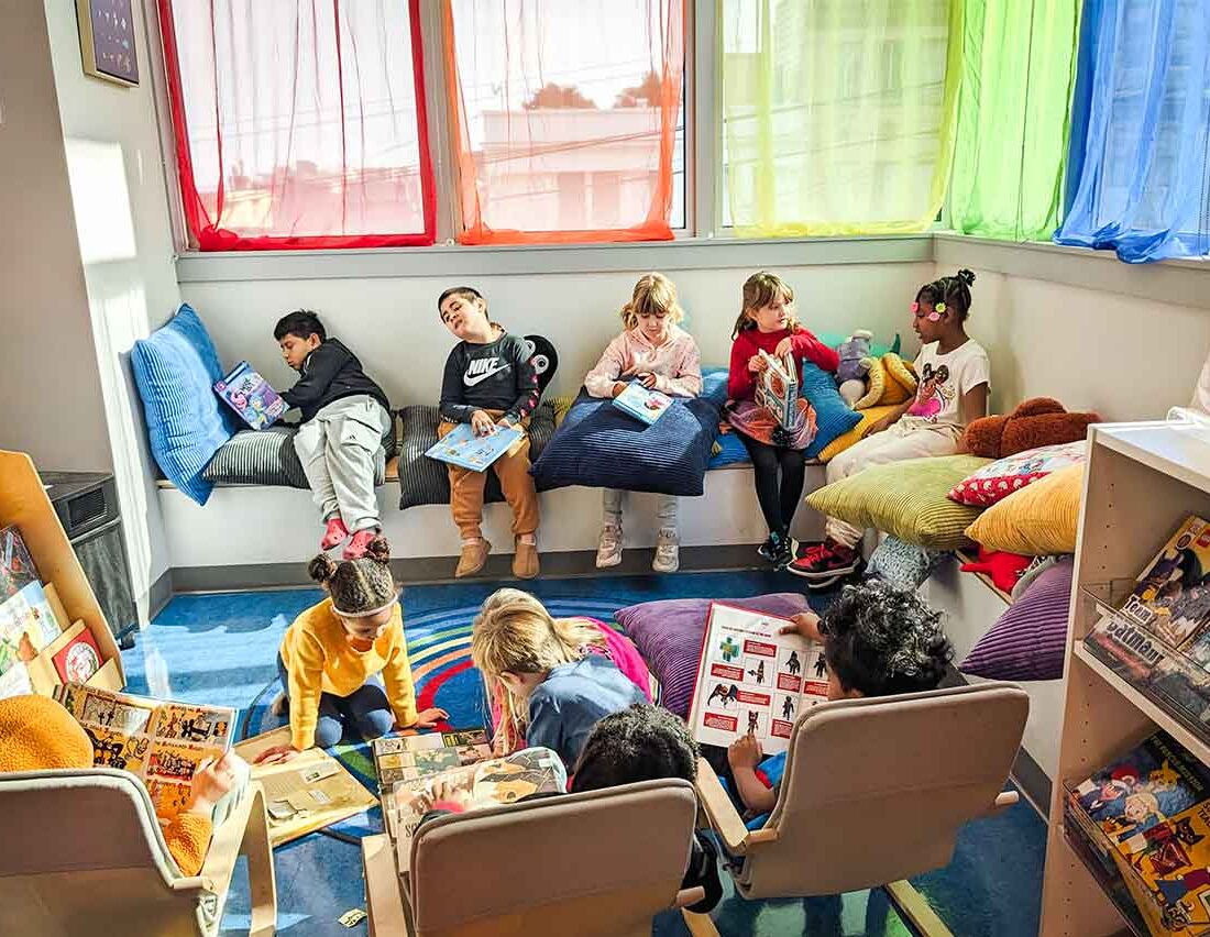 Kids sit in the library reading with rainbow colored curtains in the windows